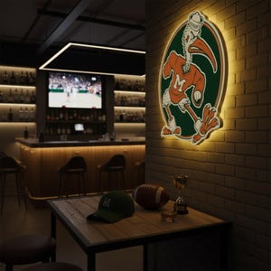 May include: A sports bar scene featuring a lit sign with a pelican mascot wearing a sailor hat. A green baseball cap with an "M" sits on a wooden table with a football, a trophy, and a glass of amber liquid. The bar has a TV and a well-stocked bar.