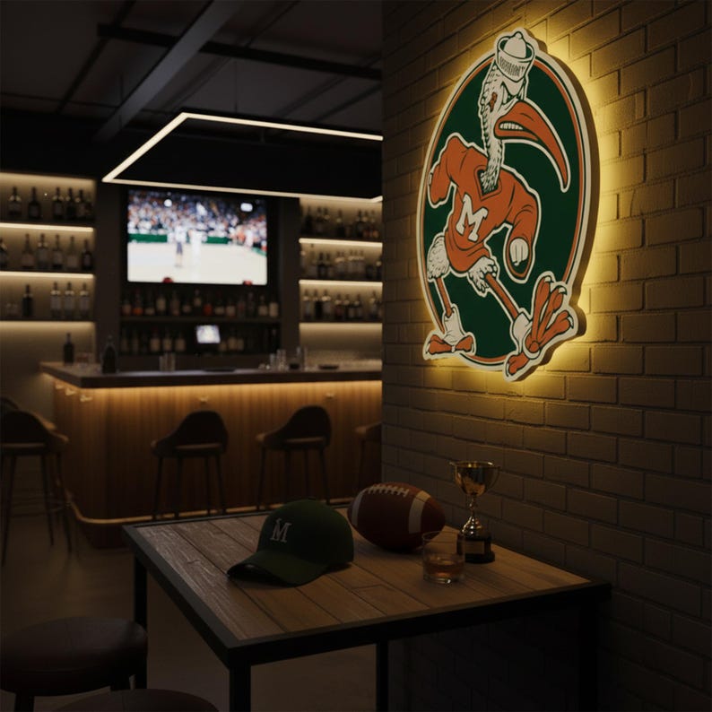 May include: A sports bar scene featuring a lit sign with a pelican mascot wearing a sailor hat. A green baseball cap with an "M" sits on a wooden table with a football, a trophy, and a glass of amber liquid. The bar has a TV and a well-stocked bar.