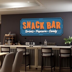May include: A blue and orange sign with the text "Snack Bar" and the words "Drinks, Popcorn, Candy" underneath. The sign is hanging on a wall above a bar counter with stools.