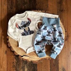 Könnte beinhalten: Ein weißes Baby-Set mit einem Cartoon-Charakter. Das langärmlige Oberteil hat eine Grafik des Charakters. Die blaue Hose hat einen passenden Charakterdruck auf hellblauem Hintergrund. Das Set wird auf einer Holzoberfläche präsentiert.