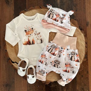 Può includere: Un set di abbigliamento per bambini con stampa di animali del bosco, tra cui un body bianco, un cappello e un bloomer. Sono incluse anche scarpine in pelle bianca. Il body presenta una grafica di volpe e scoiattolo.