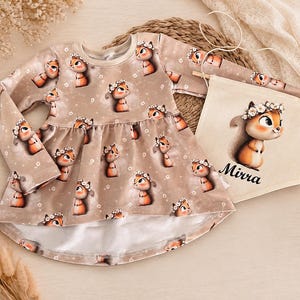 Könnte beinhalten: Ein langärmeliges Kleid in Hellbraun mit einem durchgehenden Muster aus Zeichentrick-Eichhörnchen, die Blumenkränze tragen. Neben dem Kleid liegt ein rechteckiges Stoffbanner mit einer passenden Eichhörnchen-Illustration und dem Namen Mirra.