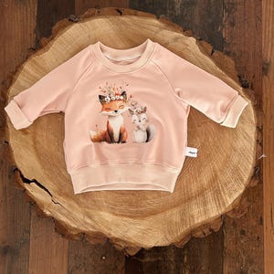 Könnte beinhalten: Ein hellrosa Sweatshirt mit Rundhalsausschnitt und langen Ärmeln. Auf der Vorderseite befindet sich eine skurrile Illustration eines Fuchses und eines Eichhörnchens, die beide mit Blumenkränzen geschmückt sind. Das Sweatshirt wird auf einer Holzoberfläche präsentiert.