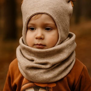 Op de afbeelding: Een kind draagt een beige hoed met oorachtige details en een bijpassende nekwarmer. Het kind draagt ook een roestkleurig shirt met lange mouwen met een cartoonpinguïn op de voorkant. De achtergrond is vervaagd met herfstkleuren.