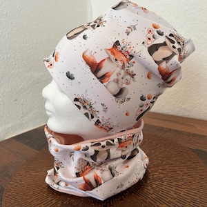 Könnte beinhalten: Ein weißes Mützen- und Halstuch-Set mit einem sich wiederholenden Muster aus Füchsen, Waschbären und floralen Akzenten. Die Füchse sind orange und weiß, und die Waschbären sind schwarz und weiß. Die floralen Akzente umfassen Orange, Rosa und Grün.