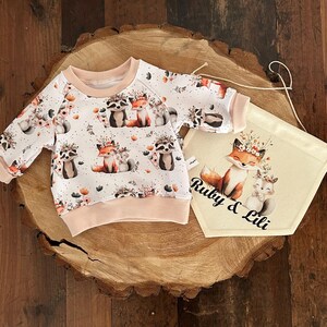 Op de afbeelding: Een witte baby-sweatshirt met een bosdierenprint met wasberen en vossen. De sweatshirt heeft een lichtroze rand en wordt gecombineerd met een bijpassende banner met de tekst "Ruby & Lili".