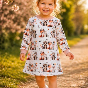 Könnte beinhalten: Ein langärmeliges Kleid mit weißem Grund und einem wiederkehrenden Muster aus Füchsen und Waschbären mit Blumenkränzen, akzentuiert durch kleine orangefarbene und graue Blüten. Das Kleid hat einen hellrosa Rundhalsausschnitt und passende Bündchen.