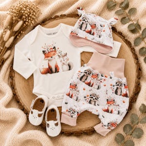 Könnte beinhalten: Ein langärmeliger weißer Body mit Fuchs- und Eichhörnchen-Motiv, eine passende Hose mit rosa Bund und Tiermuster, eine weiße Knotenmütze mit rosa Futter sowie ein Paar weiße T-Spangen-Schuhe mit ausgestanzten Details.