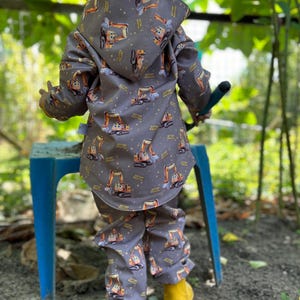 Può includere: Un completo giacca con cappuccio e pantaloni grigi per bambini piccoli, con un motivo di veicoli da cantiere a fumetti arancioni e gialli. Il bambino indossa stivali gialli. L'outfit è progettato per il gioco all'aperto.