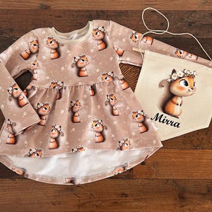 Könnte beinhalten: Hellbraunes Langarmkleid und Banner mit Cartoon-Eichhörnchen mit Blumenkränzen. Das Kleid hat einen weißen Saum. Das Banner zeigt eine Eichhörnchen-Illustration und den Namen "Mirra".