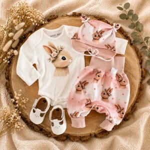Peut inclure: Un ensemble de vêtements pour bébé comprenant un body blanc à manches longues avec un motif de lapin, un pantalon rose à motif lapin répété, un bonnet rose assorti avec des oreilles de lapin et des chaussures blanches à bride en T.