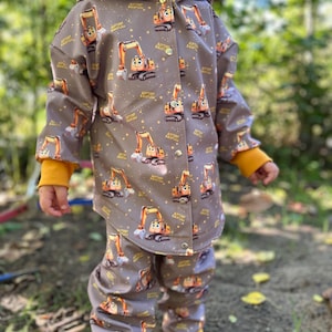 Könnte beinhalten: Grauer Regenanzug für Kinder mit einem sich wiederholenden Muster von Cartoon-Bauarbeitern. Die Jacke hat eine gelbe Kapuze und Bündchen, und die passende Hose ist ebenfalls grau. Das Kind trägt gelbe Gummistiefel.