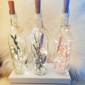 Op de afbeelding: Drie heldere glazen flessen met kurken, elk gevuld met gedroogde bloemen en feeërieke lichtjes. De flessen zijn versierd met kant en touw, en staan op een witte houten kist. De tekst "Flower Bottles" staat onderaan.