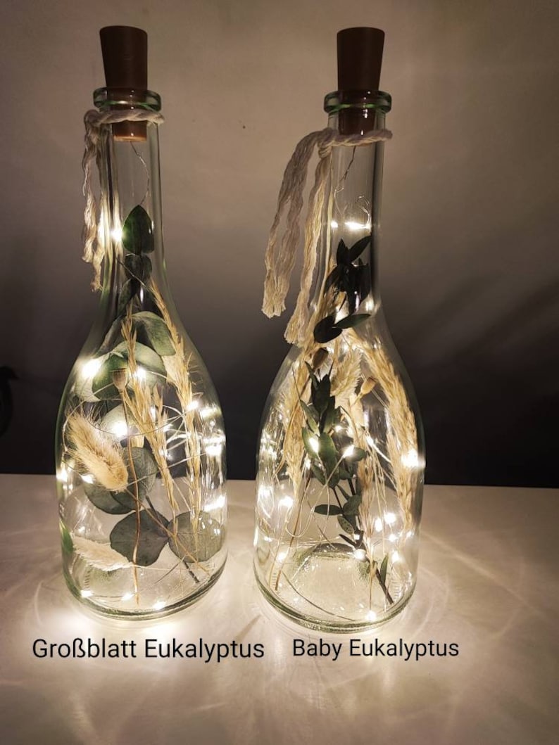 May include: Two clear glass bottles with cork stoppers, filled with dried plants and fairy lights. The bottles have a rustic aesthetic, with twine accents. The text at the bottom reads "Gro&szlig;blatt Eukalyptus" and "Baby Eukalyptus".
