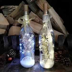 May include: Two clear glass bottles with cork stoppers, decorated with fairy lights, faux snow, and gold and silver accents. One bottle has a gold bow, the other a silver bow. The bottles are set against a backdrop of firewood.