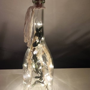 May include: A clear glass bottle with a brown cork stopper, filled with dried plants and fairy lights. A cream-colored string is tied around the neck of the bottle. The bottle is lit from within, creating a warm glow.
