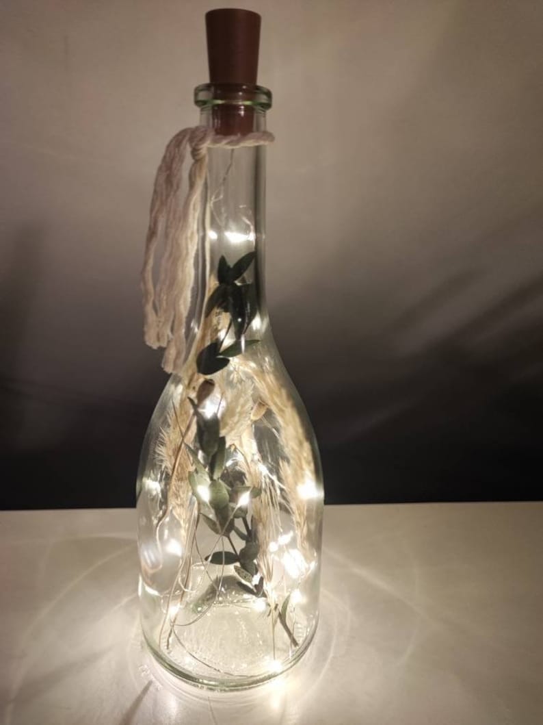 May include: A clear glass bottle with a brown cork stopper, filled with dried plants and fairy lights. A cream-colored string is tied around the neck of the bottle. The bottle is lit from within, creating a warm glow.
