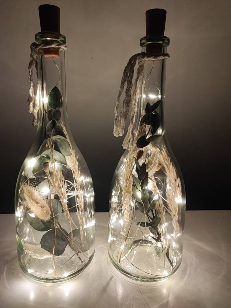 May include: Two clear glass bottles with cork stoppers, filled with dried plants and fairy lights. The bottles are decorated with twine and are illuminated, creating a warm, inviting glow. The bottles are a decorative home accent.