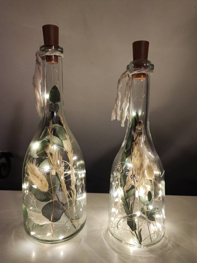 May include: Two clear glass bottles with cork stoppers, filled with dried plants and fairy lights. The bottles have a string tied around the neck. The dried plants include green leaves and beige grasses. The fairy lights are small and white.