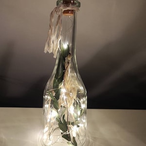 May include: A clear glass bottle with a brown cork stopper, filled with dried plants and fairy lights. The bottle is decorated with a twine bow. The lights are on, creating a warm glow. The bottle is a decorative item.