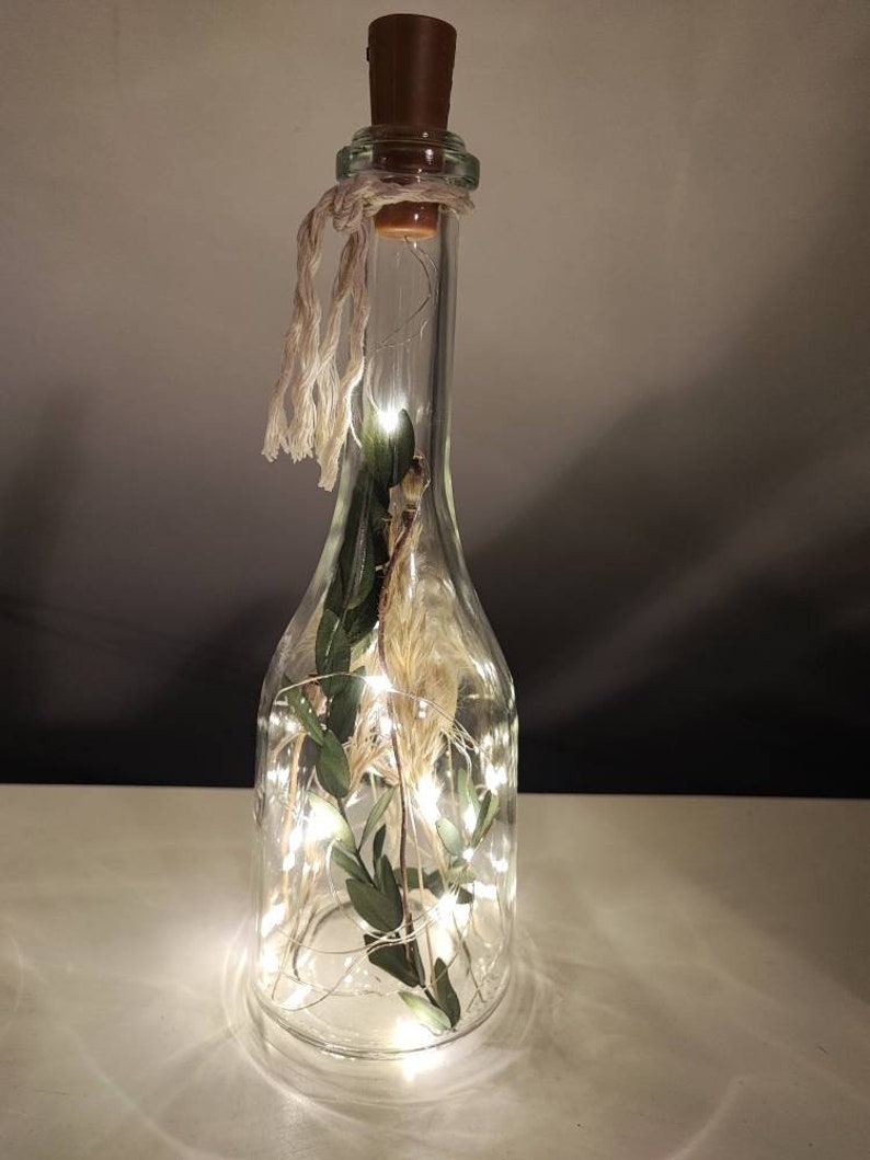 May include: A clear glass bottle with a brown cork stopper, filled with dried plants and fairy lights. The bottle is decorated with a twine bow. The lights are on, creating a warm glow. The bottle is a decorative item.
