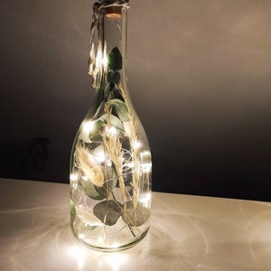 May include: A clear glass bottle with a cork stopper, filled with greenery and fairy lights. The bottle is decorated with dried plants and a string of small LED lights, creating a warm, ambient glow. A twine is wrapped around the neck of the bottle.