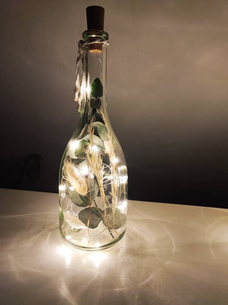 May include: A clear glass bottle with a cork stopper, filled with greenery and fairy lights. The bottle is decorated with dried plants and a string of small LED lights, creating a warm, ambient glow. A twine is wrapped around the neck of the bottle.
