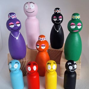 Pode incluir: Uma coleção de nove bonecas de madeira coloridas. Cada boneca é pintada com uma cor diferente, incluindo roxo, rosa, laranja, preto, verde, azul, vermelho e amarelo. As bonecas têm rostos sorridentes, algumas com tiaras florais e colares de pérolas.