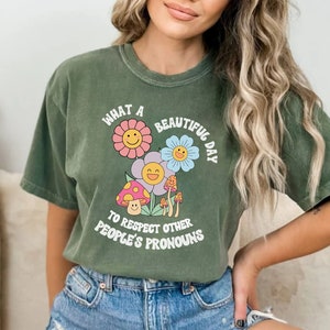 May include: A green t-shirt with a graphic of flowers and mushrooms. The text on the shirt reads "What a beautiful day to respect other people's pronouns."