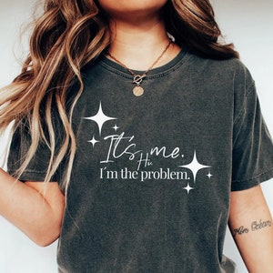 Może przedstawiać: Szary t-shirt z białym nadrukiem, kt&oacute;ry głosi "It's me. Hi. I'm the problem." z gwiazdami wok&oacute;ł tekstu.