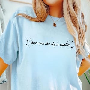 May include: Light blue t-shirt with the text "but now the sky is opalite" in black script. The text is centered and flanked by star clusters. The shirt has short sleeves and a relaxed fit.