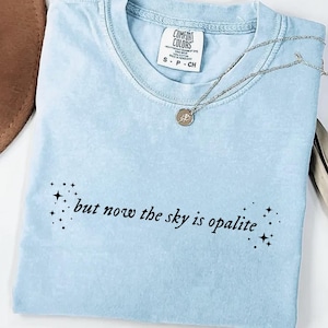 May include: Light blue Comfort Colors t-shirt with the text "but now the sky is opalite" and star graphics. A silver necklace with a pendant is draped across the shirt. The shirt is folded.