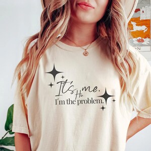 Może przedstawiać: Jasnobeżowy t-shirt z czarnym nadrukiem, kt&oacute;ry brzmi "It's me. Hi. I'm the problem." z małymi czarnymi gwiazdkami wok&oacute;ł tekstu.