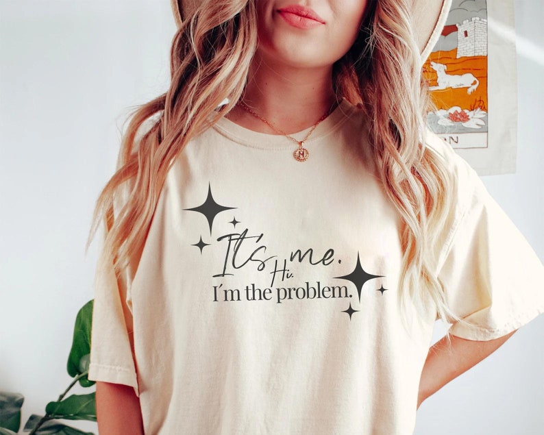 Może przedstawiać: Jasnobeżowy t-shirt z czarnym nadrukiem, kt&oacute;ry brzmi "It's me. Hi. I'm the problem." z małymi czarnymi gwiazdkami wok&oacute;ł tekstu.