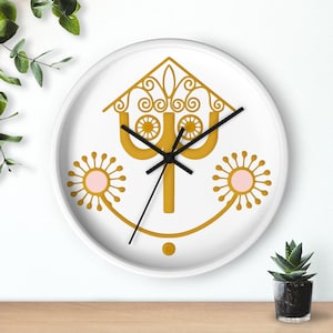 Puede incluir: Reloj de pared blanco con un diseño caprichoso de color dorado y manecillas negras. El reloj presenta una forma de casa estilizada con remolinos decorativos, dos formas de sol con centros rosas y una cara sonriente.