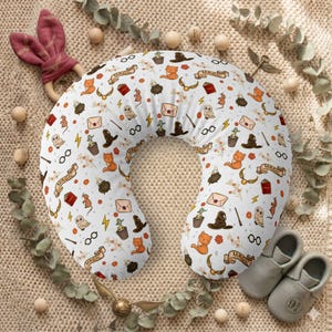 May include: A white U-shaped nursing pillow featuring a repeating pattern of orange cats, owls, black cauldrons, brown sorting hats, golden winged balls, eyeglasses, wands, envelopes, and scrolls with the word MAGIC printed in black ink.
