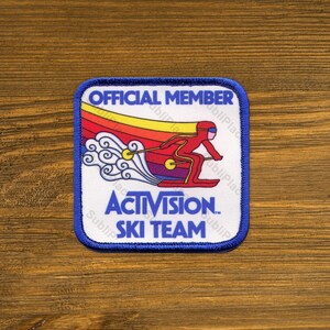 May include: A square patch with a blue border and the words "OFFICIAL MEMBER" at the top. The patch features a skier in red and a rainbow design, with the words "ACTIVISION SKI TEAM" below. The patch is on a wooden surface.