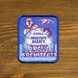 May include: A rectangular embroidered patch with a blue border featuring an illustration of an igloo, a polar bear, a bird, and a crab. The text reads Activision, Frostbite Bailey's, and Arctic Architects in blue and white lettering.