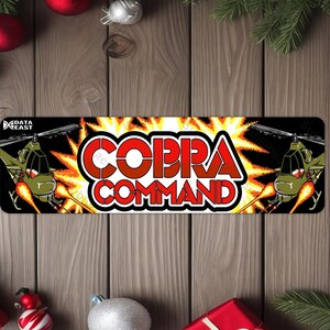 May include: A rectangular sign with the words "COBRA COMMAND" in red and white, with a yellow and orange explosion background. Two green helicopters with flames are on either side of the text. The sign has the "DATA EAST" logo in the upper left corner.