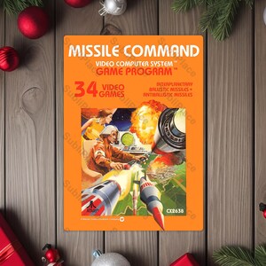 May include: An orange Missile Command video game cartridge. The cover art features a retro illustration of a missile launch with the text "MISSILE COMMAND" and "34 VIDEO GAMES". The Atari logo is visible.