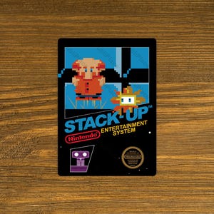 May include: A black sticker featuring pixelated graphics from the Nintendo game "Stack Up." The design includes a character in an orange coat, the game's logo, and the Nintendo logo. The sticker is rectangular.