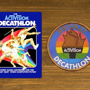 May include: A blue and yellow video game cartridge for the Atari 2600 video computer system. The cartridge features the text "The Activision. Decathlon" and a graphic of athletes competing in a decathlon. A bronze medal patch with the text "Bronze Decathlon" and the Activision logo is also shown.