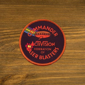 May include: A purple and orange embroidered patch with the text "Commander of the Federation of Laser Blasters" and a cartoon spaceship. The text "Activision" is in the center of the patch.