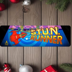 May include: A rectangular mousepad featuring the Atari game "S.T.U.N. Runner." The design includes a red spaceship, a cityscape, and the game's logo in yellow and orange. The mousepad is set against a blue and black background.