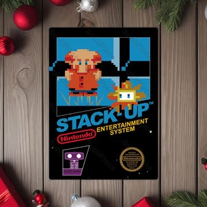 May include: A black Nintendo Entertainment System game cartridge featuring pixelated characters and the game title "Stack Up." The cartridge has a retro design with the Nintendo logo and a gold seal. The background includes Christmas decorations.