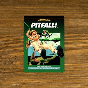 May include: A green and brown video game cartridge for the Intellivision console. The front of the cartridge features a cartoon image of a man swinging on a vine, with a snake and jungle foliage in the background. The text "ACTIVISION. PITFALL!" is printed in white on a red background.