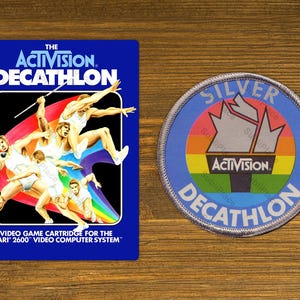 May include: A blue and rainbow colored video game cartridge for the Atari 2600 video computer system. The cartridge is labeled "The Activision. Decathlon". A silver patch with the same text and a rainbow design is also shown.