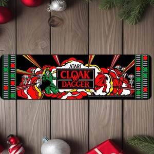 May include: A black and red arcade game cabinet with the text "ATARI CLOAK & DAGGER" on the front. The cabinet has a green and red border with a repeating pattern.