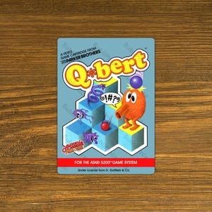May include: A vintage Atari 5200 game cartridge featuring the game "Q*bert." The cartridge has a light blue background with the game's title in yellow and a cartoon character. The cartridge is from Parker Brothers.
