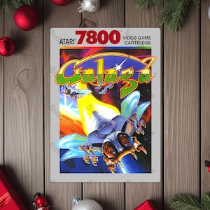 May include: Atari 7800 video game cartridge for Galaga. The cartridge features a colorful illustration of a spaceship with blue wings and a yellow beam. The Atari logo and game title are displayed at the top.